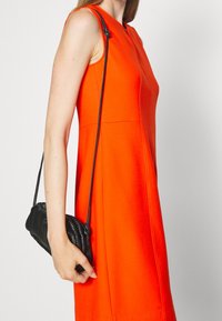 Robe orange ajustée avec un design sans coutures et un sac à main noir texturé avec une longue bandoulière, tenu dans la main droite du mannequin.