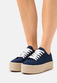 Námořnické plátěné tenisky na platformě s bílými tkaničkami, jute rope podrážkami a kovovými očky; zaoblená špička a minimální detaily šití.