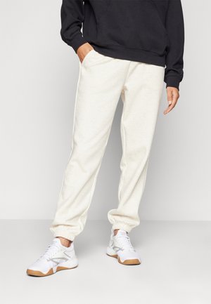 Weiße Fleece-Jogginghosen mit elastischen Bündchen, kombiniert mit einem schwarzen Sweatshirt. Das Model trägt weiße Sportschuhe mit braunen Sohlen.