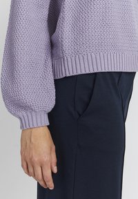 Helllila gestrickter Pullover mit strukturiertem Muster und gerippten Bündchen, kombiniert mit dunkelblauen, taillierten Hosen.