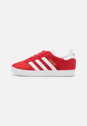 Zapatilla Adidas Gazelle roja con rayas blancas y suela blanca, textura de ante, y texto dorado "GAZELLE" en el lateral.