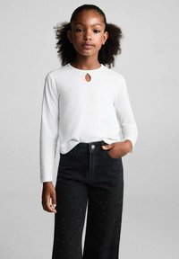 Bílý dlouhý rukáv top s detaily ve tvaru klíčové díry na výstřihu, doplněný o černé džíny s vysokým pasem a jemnými ozdobami.