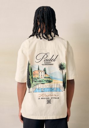 Persoon met gevlochten haar die een wit shirt draagt en op de achterkant een kleurrijke padel tennisbaan en Italiaanse landschapsillustratie toont.