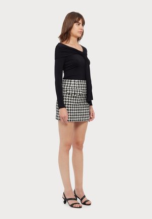 Musta pitkähihainen paita, jossa on roikkuva kangas, yhdistettynä korkeavyötäröiseen musta-valkoiseen houndstooth-minihameeseen ja mustiin remmisandaaleihin.