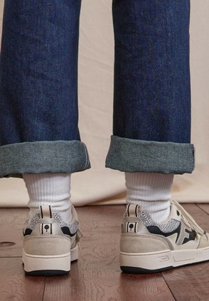 Personne portant un jean bleu foncé retroussé, des chaussettes blanches côtelées et des baskets beige avec des accents noirs et à motifs, debout sur un sol en bois.