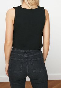 Frau in einem ärmellosen schwarzen Oberteil und hoch geschnittenen dunkelgrauen Jeans, von hinten gezeigt, mit einem sichtbaren Handtuch-Tattoo am rechten Handgelenk.