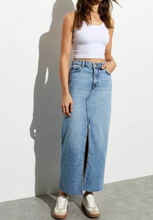 Femme portant un crop top blanc sans manches, une longue jupe en jean bleu clair taille haute avec une fente devant, et des baskets blanches beiges, debout contre un mur blanc.