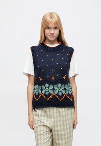 Granatowy bezrękawnik z dzianiny z pomarańczowo-zielonymi wzorami kwiatowymi, z fakturowaną powierzchnią i prążkowanym dekoltem. Noszony na białej koszuli.