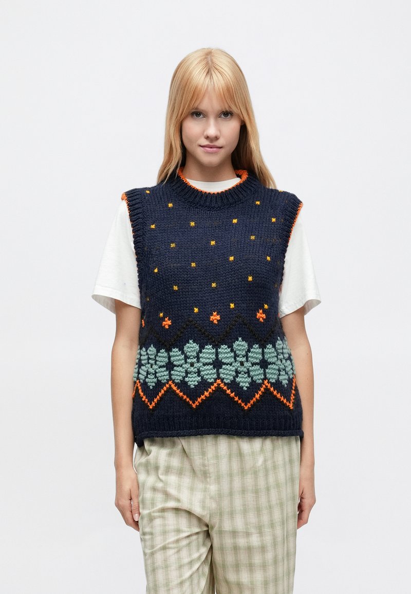 Granatowy bezrękawnik z dzianiny z pomarańczowo-zielonymi wzorami kwiatowymi, z fakturowaną powierzchnią i prążkowanym dekoltem. Noszony na białej koszuli.