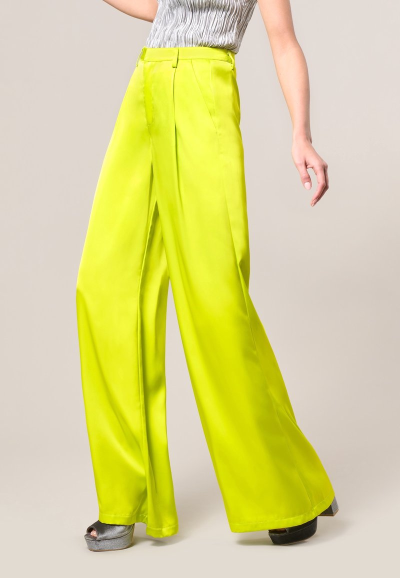 Jasno-limonkowe, szerokie spodnie z gładkiego materiału, z wysokim stanem i dwoma fałdami z przodu, w zestawieniu z srebrnymi sandałami na platformie.