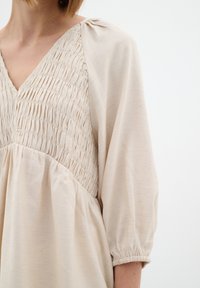Vestito beige chiaro con scollatura a V, caratterizzato da arricciature sul busto e maniche corte a sbuffo. Tessuto morbido e strutturato con una leggera lucentezza.