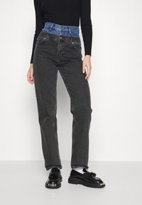 Vaqueros de mezclilla negros de talle alto con una cinturilla de mezclilla azul, que cuentan con cinco bolsillos, una cremallera frontal y un diseño de pierna recta. Zapatos negros brillantes.