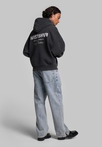 Felpa nera oversize con testo bianco sul retro, abbinata a jeans in denim azzurro chiaro a vestibilità ampia e scarpe nere. Tessuto morbido.