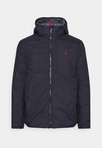 Mörkblå puffjacka med huva, som har en framfällbar dragkedja, röda detaljer och en liten broderad logotyp på bröstet. Slät, vattenavvisande material.