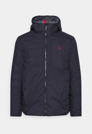 Mörkblå puffjacka med huva, som har en framfällbar dragkedja, röda detaljer och en liten broderad logotyp på bröstet. Slät, vattenavvisande material.