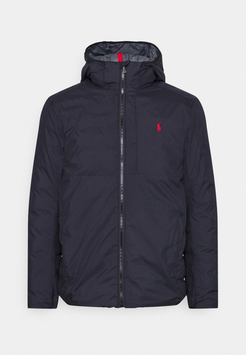 Mörkblå puffjacka med huva, som har en framfällbar dragkedja, röda detaljer och en liten broderad logotyp på bröstet. Slät, vattenavvisande material.