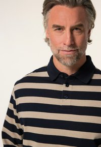 Granatowa i beżowa w paski koszulka polo z kołnierzykiem, z trzema guzikami i miękką bawełnianą strukturą. Zbliżenie na górną część ciała.