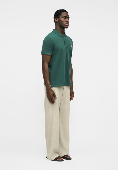 Homme portant un polo à manches courtes vert foncé, un pantalon beige à jambes larges, et des sandales noires, debout devant un fond blanc uni.