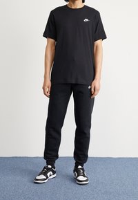 Svart Nike T-shirt med rund hals och logotyp, ihop med svarta sweatpants med en logotyp på låret, bärs med svarta och vita sneakers.