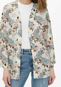 Giacca kimono floreale in tessuto crema. Presenta motivi floreali blu e marroni, apertura frontale e maniche larghe. Abbinate a una maglietta bianca e jeans.