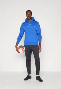 Sweatshirt azul com bolsos frontais e cordão, combinado com calças de jogging pretas e ajustadas. O modelo segura uma bola de futebol e usa tênis esportivos pretos.