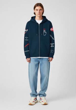 Katoenen blend zip-up hoodie in donkerblauw met grafische prints en tekst op de mouwen, met een kangoeroezak en een capuchon met trekkoord. Draagt met lichtblauwe jeans.