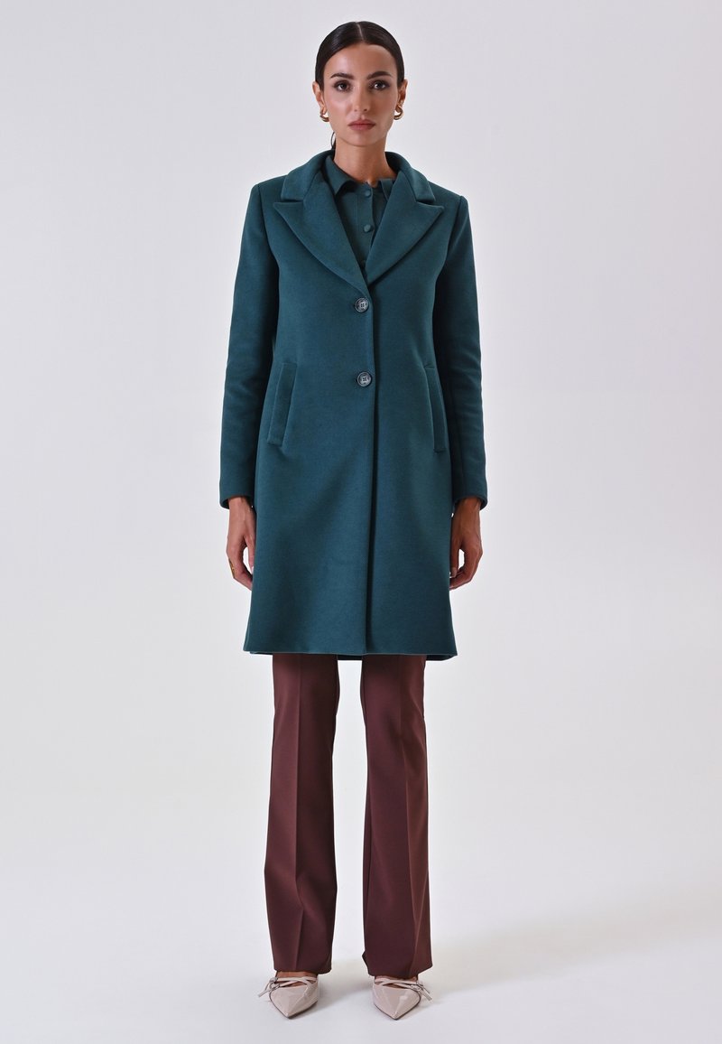 Cappotto in lana teal con colletto a punta, due tasche anteriori, tre bottoni, abbinato a pantaloni marroni e scarpe chiare.