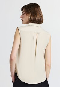 Blusa beige senza maniche con colletto a bottone, dettaglio di piega sul retro e orlo arrotondato. Realizzata in un tessuto liscio con una leggera texture.