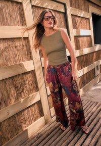 Débardeur kaki texturé associé à un pantalon large bordeaux à motif floral. Le modèle porte des lunettes de soleil et des sandales noires à brides. Fond en bois.