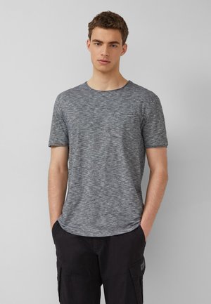 Junger Mann mit kurzen lockigen Haaren, der ein graues T-Shirt mit kurzen Ärmeln und Taschen sowie eine schwarze Cargohose trägt, steht vor einem einfarbigen Hintergrund.