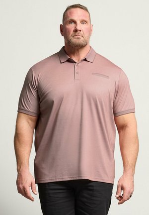 Homme portant un polo mauve à manches courtes et un pantalon noir, debout sur un fond clair uni, face à l'avant.
