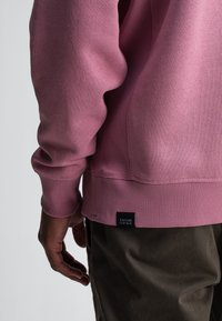 Rosa Sweatshirt mit gerippten Bündchen und Saum, glatter Textur und einem schwarzen Etikett mit "BUTCHER OF BLUE" an der unteren Seite.
