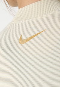 Tkanina w kolorze kremowym z poziomymi paskami, z złotym logo Nike na ramieniu. Gładka tekstura i dopasowany krój.
