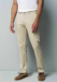 Beige chino's met een slim fit, platte voorkant en subtiele glans. Voorzien van een knoopsluiting en zijzakken, gecombineerd met bruine suède schoenen.