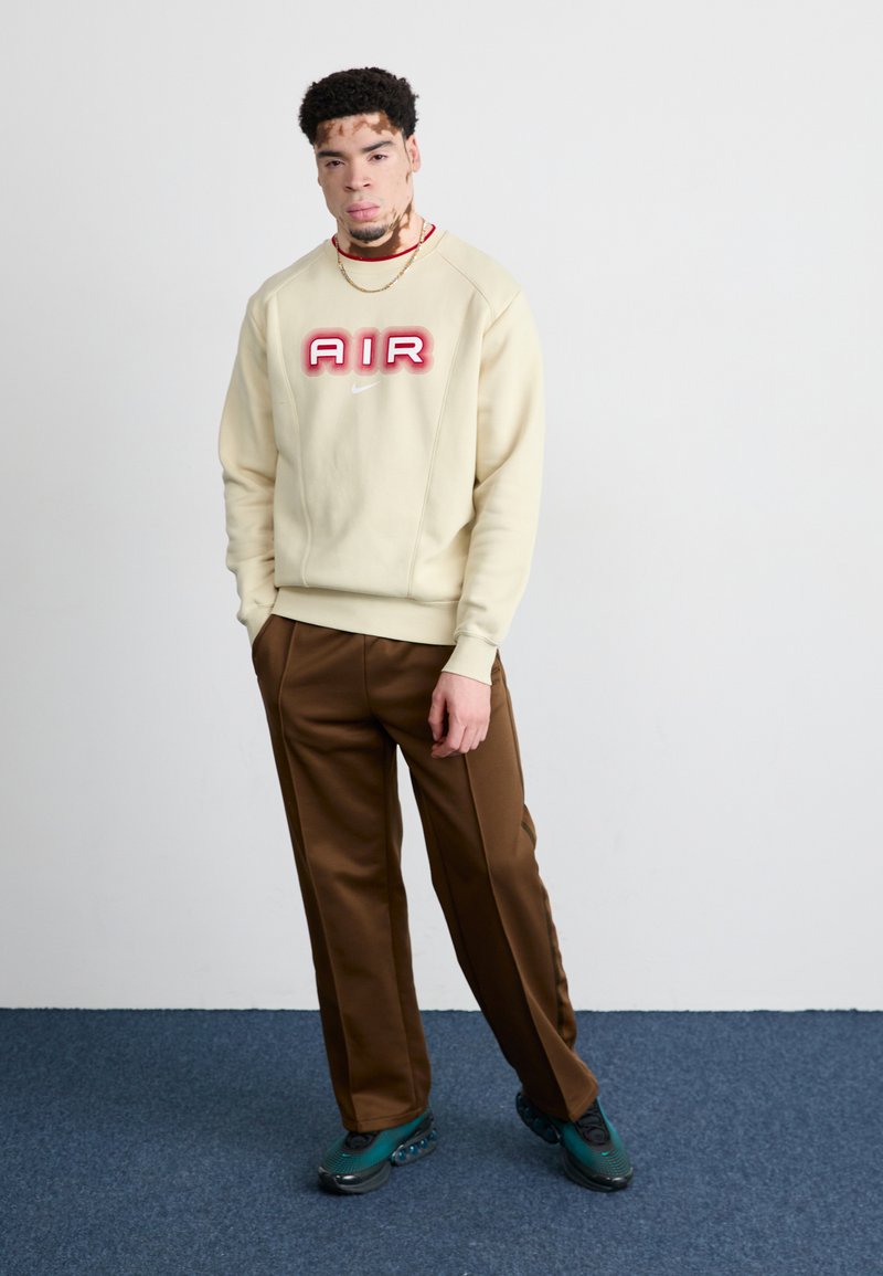 Cremefarbener Sweatshirt mit rotem "AIR"-Logo, kombiniert mit braunen maßgeschneiderten Hosen. Das Outfit wird mit dunkelgrünen Sneakern mit strukturiierter Sohle abgerundet.