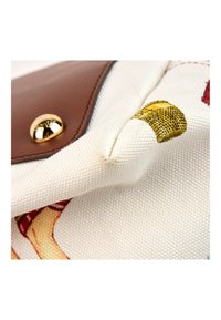 Gros plan d’un coin de sac en toile beige avec un rabat en cuir marron et un fermoir bouton doré, avec un petit détail imprimé jaune.