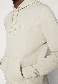 Beige hoodie med en framficka, dragsko huva och en liten logotypapplikation. Tillverkad av mjukt tyg med synliga sömdetaljer.