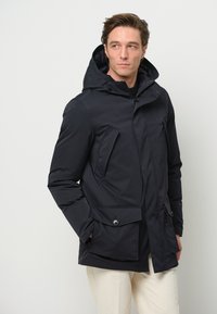 Marinblå pufferjacka med huva, med dragkedja och knäppning, två sidofickor och en slät, mjuk tygstruktur.