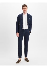 Marineblazer mit zwei Knöpfen, passende maßgeschneiderte Hose und ein weißes Hemd. Braune Slipper runden das Outfit ab. Glattes Material und taillierte Passform.