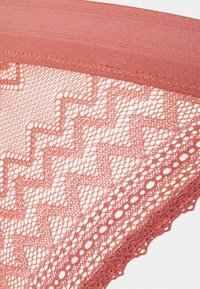 Lingerie en dentelle de couleur rose poussiéreux, avec un bord festonné, des motifs géométriques et un ceinturon lisse. Les détails en dentelle texturée sont bien visibles.