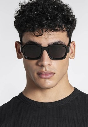 Jeune homme aux cheveux noirs bouclés portant de grandes lunettes de soleil rectangulaires noires et un t-shirt noir à col rond côtelé contre un fond uni.