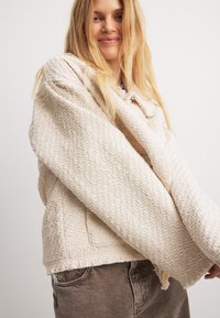 Beige texturerad jacka med lös passform, mönstrad tyg och breda ärmar; har en knappstängning och fransade fållar.