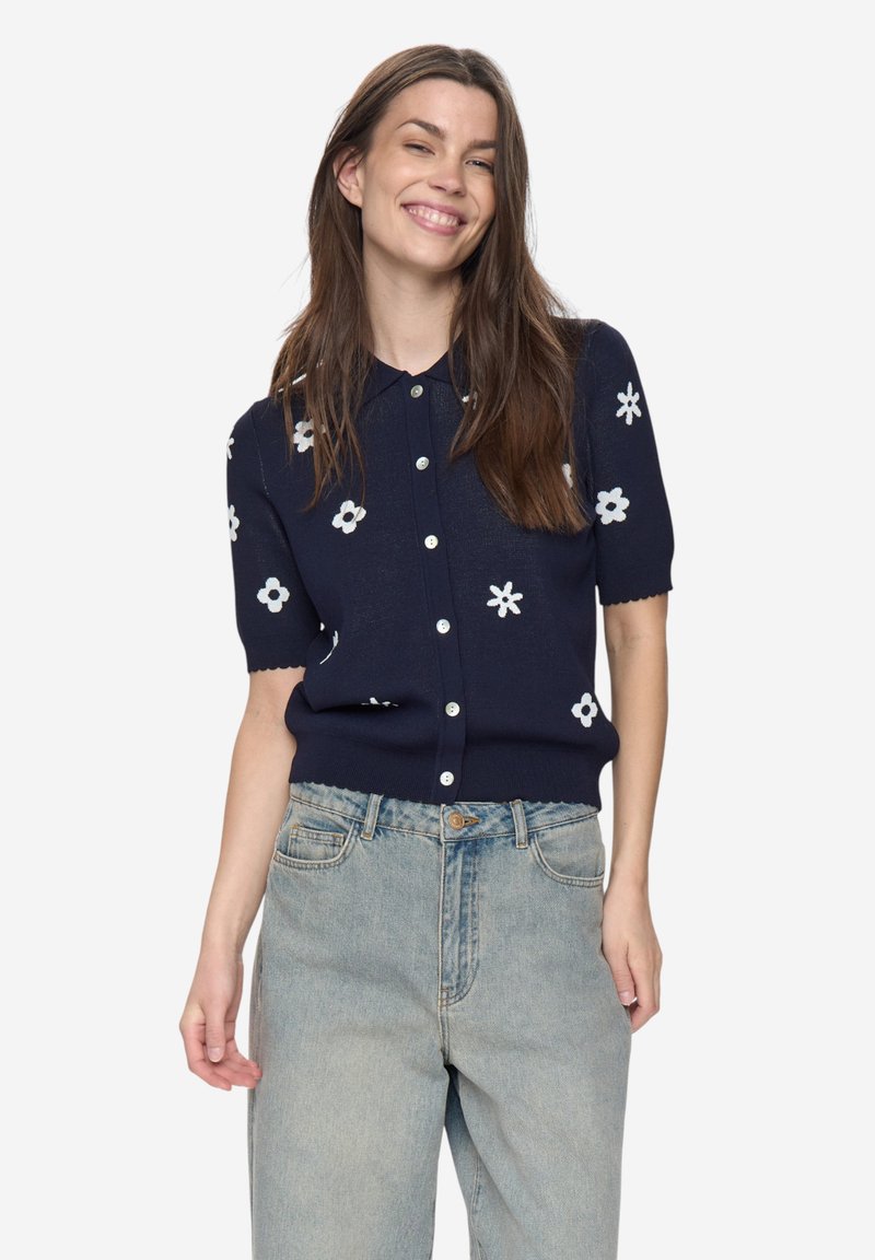 Marineblauwe gebreide korte mouw cardigan met witte bloemenpatronen en knopen aan de voorkant. Voorzien van geschulpte randen aan mouwen en zoom, gemaakt van zacht materiaal.