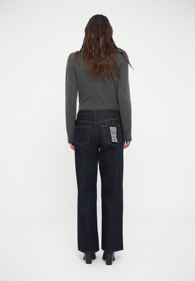 Jeans a gamba larga blu scuro in denim, con un delicato motivo a cucitura sulla tasca posteriore. Abbinati a un maglione grigio.