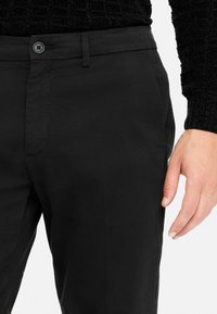 Close-up of a person wearing black pants and a black textured sweater, with their right hand relaxed by their side.