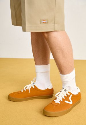Jambes portant un short beige, des chaussettes blanches hautes et des baskets basses orange avec des lacets blancs et une semelle en caoutchouc, debout sur un sol moutarde.