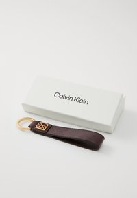 Porte-clés en cuir marron Calvin Klein avec un anneau en métal doré et un accent gravé du logo, présenté dans une boîte blanche.