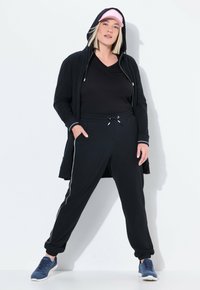 Schwarzes Sportoutfit bestehend aus einer zipfbaren Jacke mit Kapuze, lockerer Passform Hose mit seitlichen Streifen und marineblauen Sneakers. Akzentuiert mit einer pinken Kappe.