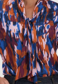 Multikleurige blouse met een mix van blauwe, oranje en roze abstracte patronen. Zachte stof met een strik bij de hals en een vloeiende design.