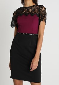 Enges Kleid mit einem Pflaumenoberteil und schwarzem Spitzenoverlay, das einen gewellten Ausschnitt aufweist. Ein schwarzer Stoffgürtel sitzt an der Taille. Strukturiertes Spitzen-Detail.